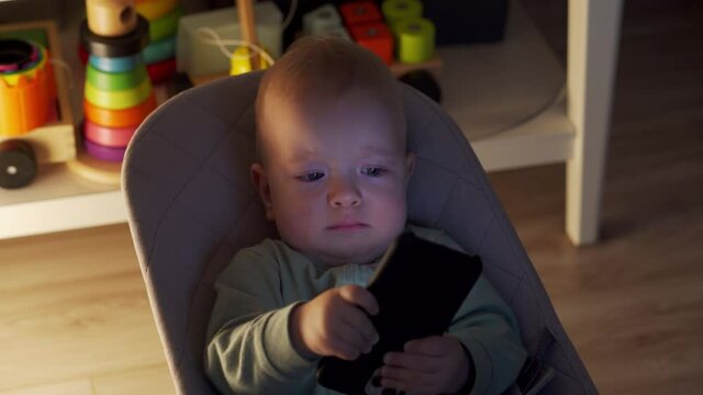 Cute toddler boy sitting on rocking chair in living room and watching something on mobile phone screen, 9 months old caucasian baby using smartphone at home. High quality 4k footage