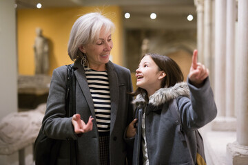 Inquisitive preteen girl visiting museum of ancient sculpture with her grandmother, exploring...