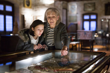 Positive senior woman and emotional inquisitive tween girl observing with interest arts and crafts...