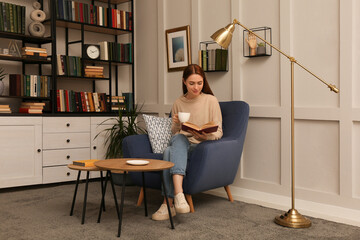 Young woman with cup of coffee reading book in armchair indoors. Home library