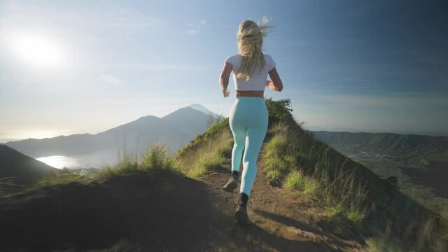 Fit sportive woman running on ridge of mount Batur towards edge, raising arms into air, high on life, freedom