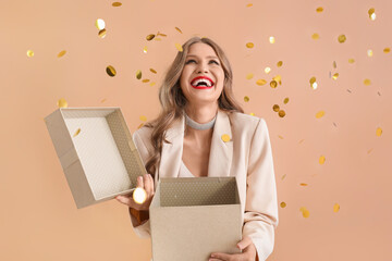 Beautiful young woman with gift and falling confetti on beige background - obrazy, fototapety, plakaty