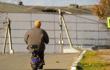 An elderly man rides along the road on a scooter