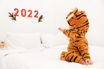 Cute caucasian baby,toddler, child, kid about 1-2 year old wearing tiger costume on white bed indoors pointing,looking at numbers 2022,back view.Symbol Chinese calendar New Year.Festive,greeting card.