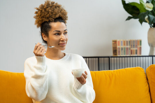 Beautiful Black Woman Eating Yogurt As Healthy Breakfast Or Snack At Home Sitting On The Couch