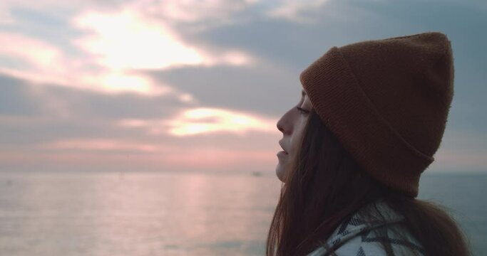 The profile of a female wearing a bonnet and looking at the sea at sunset. She sighs and turns her head slightly.