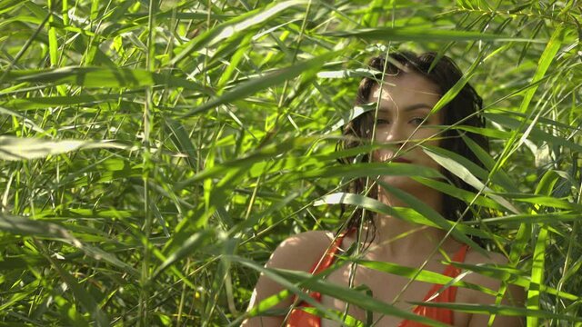 A young woman in the bulrush looks at the camera. Natural surroundings outside. The woman's face is lit by the sun's rays in the tall grass. Slowmotion movie.