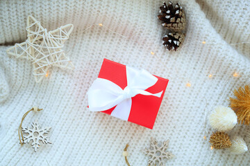 The red gift box of bokeh in the house on the table with a sweater in the background and decorations. Winter mood, festive decoration, magical Christmas.