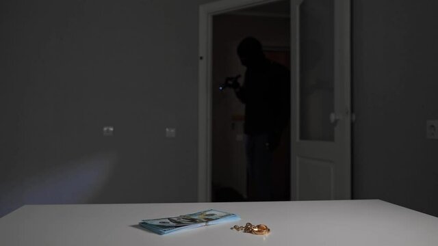 Burglar looser under disguise holding illuminating flashlight goes past unnoticed money and gold jewelry on white wooden desk in dark room closeup.