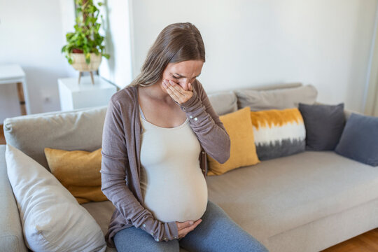 young pregnant woman suffering from toxicosis at home. maternity lady having morning sickness covering mouth with hands while sitting on couch in living room. mom expect unborn baby