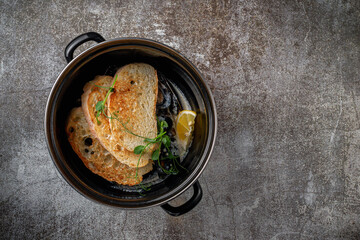 Serving a dish from a restaurant menu. Fried mussels with croutons and lemon in a dark pan against a gray stone table