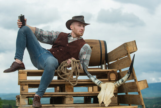 Cowboy farmer man in country side wearing western cowboy hat. Guy drinking whiskey. American Male model in countryside on farm.