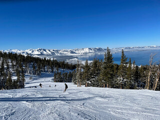 Scenic view of skiers and snowboarders on the slopes of a ski resort on a bluebird winter day, with Lake Tahoe in the background - obrazy, fototapety, plakaty