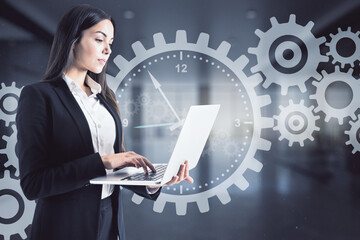 Attractive young european businesswoman using laptop with abstract cogwheel hologram on blurry office interior background. Industry and time management concept. Double exposure.