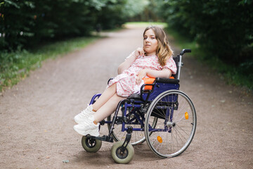 Fototapeta na wymiar Portrait of young caucasian woman with disability in stylish summer dress looking at camera with green bushes on background. Time spending in fresh air. Lifestyles of wheelchair user.