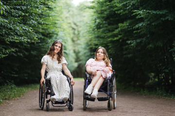 Fototapeta na wymiar Two caucasian women in wheelchairs posing on camera among green city park. Female friends who live with disability having hairstyle and wearing beautiful summer dresses.
