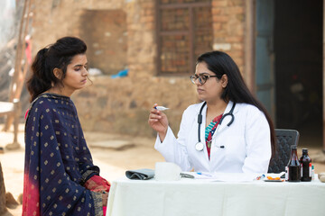 Indian female doctor holding digital thermometer checking young adult girl patient at village,...