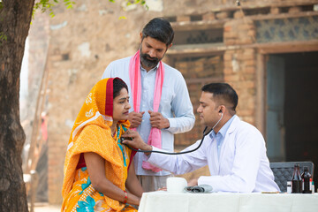 Indian male doctor with stethoscope checking patient heart beat or breath at village, woman wearing...