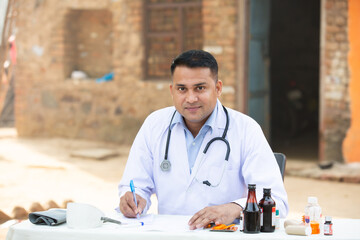 Portrait Of Young Indian Male Doctor writing prescription on paper while sitting at desk outdoor...