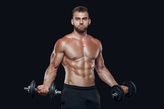 Sexy athletic man is showing muscular body with dumbbells standing with his head down, isolated over black background