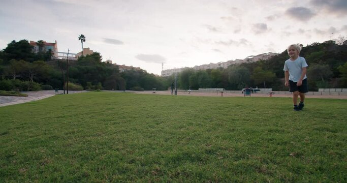 Camera tracking a happy little boy in a white T-shirt running across the green lawn playing ball kicks it towards the camera in the sunset light. Slow motion kid football in city park.