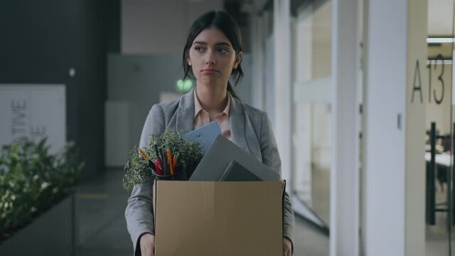 End of career. Young fired upset woman walking through office building, carrying box with personal belongings and crying