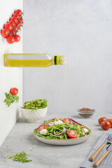 Olive oil pouring from glass bottle into vegetarian salad made of cherry tomatoes, rocket leaf or arugula, mozzarella cheese and onion rings served on plate with fork on gray concrete table