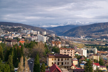 View of the city from a height against the background of snow-covered mountains and cloudy sky. Beautiful panorama of Sochi in winter.