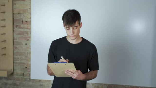 Male student presentation in classroom. Dark hair talking smiling and wearing winter clothes. Standing with the notebook in the modern classroom near the whiteboard and writing.