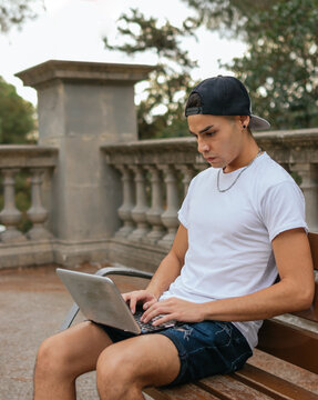 A vertical shot of a guy with cap studiying with his laptop on a public bench of a town square