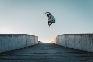 Young caucasian man in jeans and hoodie jumping on concrete bridge. Mid air parkour pose in city environment and clear sky - obrazy, fototapety, plakaty