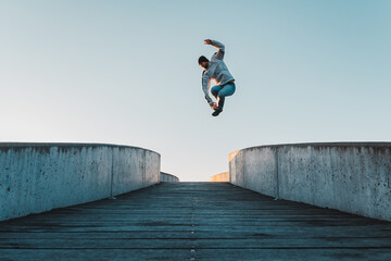 Young caucasian man in jeans and hoodie jumping on concrete bridge. Mid air parkour pose in city environment and clear sky - 476195065
