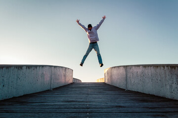 Young caucasian man in jeans and hoodie jumping with spread out arms on concrete bridge. Mid air parkour pose in city environment and clear sky - obrazy, fototapety, plakaty