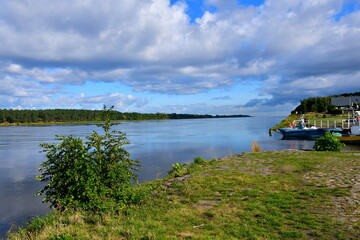 Fototapeta premium A view of a small passenger boat standing in the dock located next to the bank of a river or lake surrounded with forests and moors spotted on a cloudy summer day right before a strong thunderstorm