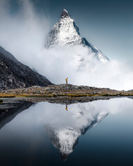Hiker man walks alone through the incredible Swiss Alps with the Matterhorn in the background between clouds and mystery while it is reflected in the Riffelsee lake - obrazy, fototapety, plakaty