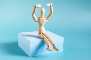 A mannequin of a man made of wood with his hands raised above his head, sitting on a blue podium close-up.
