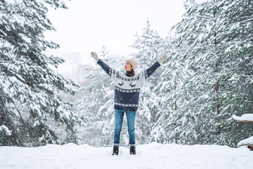 A young woman in a winter forest stands with her eyes closed and her hands raised up. Around the pine trees, covered with white snow. A frosty winter morning.