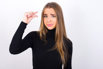 Upset Young caucasian girl wearing black turtleneck over white background shapes little gesture...