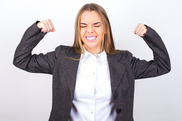 Strong powerful Young business woman wearing jacket over white background toothy smile, raises arms and shows biceps. Look at my muscles!