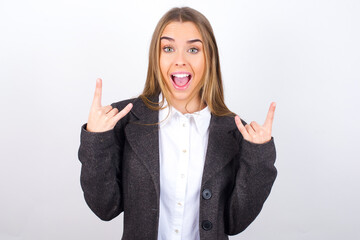 Young business woman wearing jacket over white background makes rock n roll sign looks self...