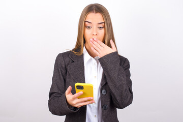 Young business woman wearing jacket over white background being deeply surprised, stares at smartphone display, reads shocking news on website, Omg, its horrible!