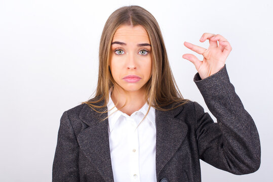 Young business woman wearing jacket over white background purses lip and gestures with hand, shows something very little.