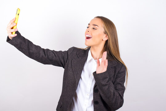 Young business woman wearing jacket over white background holds modern mobile phone and makes video call waves palm in hello gesture. People modern technology concept