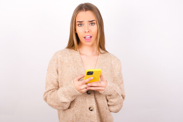 Photo of astonished crazy young caucasian girl wearing knitted sweater over white background hold smartphone dislike feedback concept