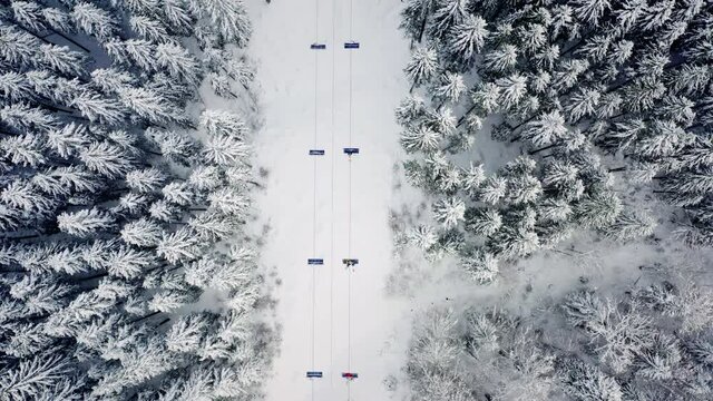 Alpine ski lift aerial drone view. Clairlift at ski resort, mountain winter forest top down rising shot. Pine trees covered with snow. Skiers and snowboarders carried up the hill. Active lifestyle.