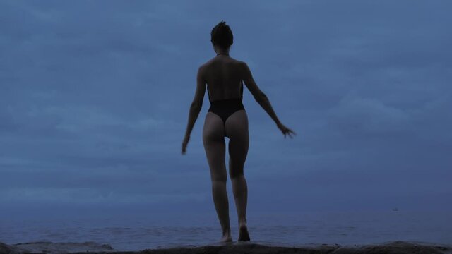 Back view of slender happy women in body suite on ocean beach on blue sunset.