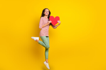 Photo of funny cute mature lady dressed striped shirt jumping high holding big red heart smiling isolated yellow color background