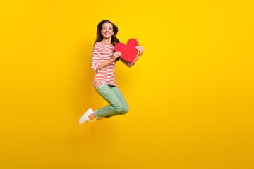 Photo of charming pretty young woman wear striped shirt jumping high holding large heart empty space isolated yellow color background