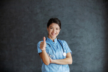 Smiling nurse with stethoscope around neck looking at camera and showing thumbs up. Health is good.
