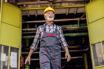 A happy senior factory worker exiting the factory and holding a walkie-talkie in his hands. A...